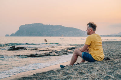 Side view of woman sitting on beach against sky during sunset