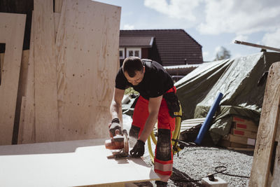 Male carpenter cutting wooden plank from electric saw while working at site during sunny day