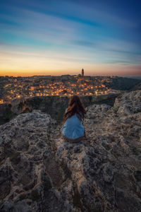 Rear view of woman sitting on rock looking at city against sky