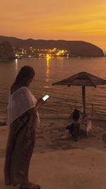 Rear view of woman standing at beach against sky during sunset reading a cellphone