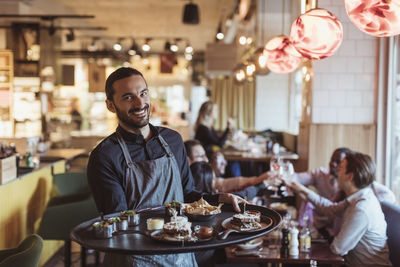 Portrait of male waiter with food tray while customers toasting in background at bar