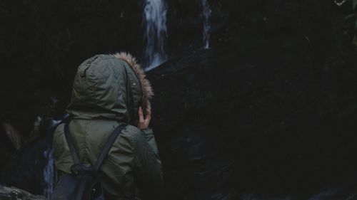Rear view of woman in warm clothing standing against waterfall