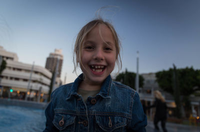 Portrait of cheerful girl against pond in city during dusk