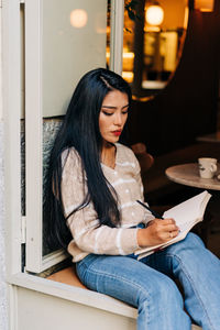 Young ethnic female in casual wear taking notes in diary while sitting on cafeteria windowsill