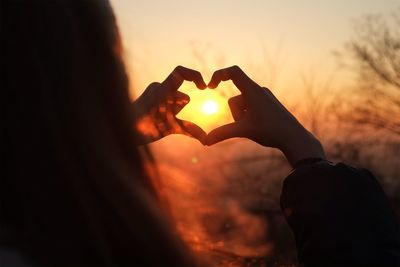 Cropped image of woman hand making heart shape against sky during sunset