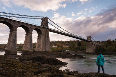 Rear view of man looking at bridge while standing by river against sky