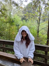 Portrait of young woman wearing white warm clothing while standing on footbridge in forest