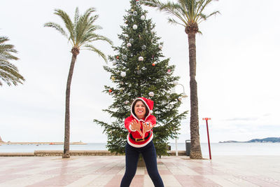 Portrait of young woman standing by palm tree against sky