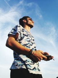 Low angle view of man holding smart phone and cigarette while standing against sky