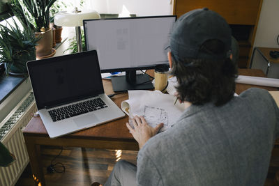 Rear view of male architect drawing blueprint on paper at desk in home office