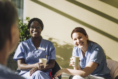 Smiling female medical workers holding coffee glasses and talking with colleague at hospital