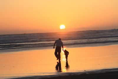 Rear view of father with son walking at beach against orange sky