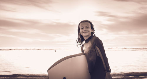 Portrait of smiling girl standing with surfboard at beach against sky during sunset