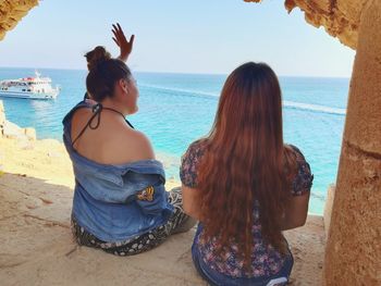 Rear view of friends looking at sea while sitting on rock formation against sky