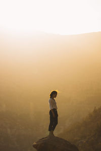 Side view of woman standing on rock against sky during sunset