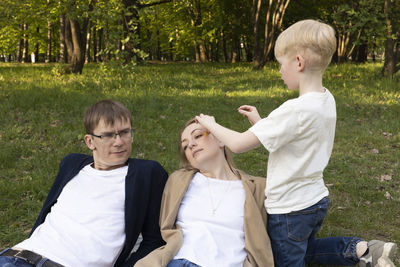 Cute caucasian 7 yo boy puts, inserts flower in mother's hair in park. mom and dad lying on grass