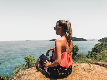 Woman sitting on cliff by sea against clear sky