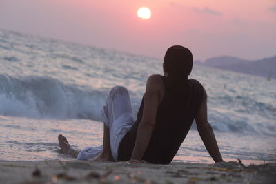 Man sitting at beach against sky during sunset