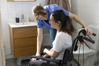 Happy female caregiver talking to woman with paraplegia in bathroom at home