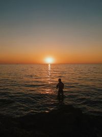 Silhouette person standing in sea against sky during sunset