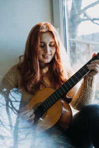 Unaltered candid portrait of young red haired woman in sweater playing acoustic guitar sitting by