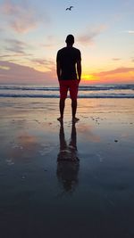 Rear view of young man standing on beach against sky during sunset