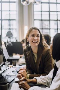 Happy mid adult businesswoman discussing with female colleague sitting at desk in office