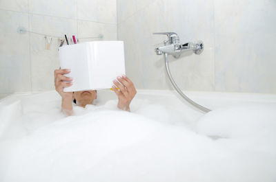 Woman lying in bathtub while reading book