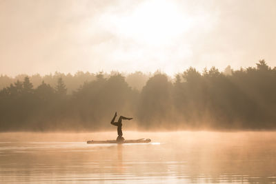 Silhouette person doing handstand in lake by trees against sky