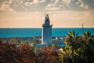 Lighthouse by sea and buildings against sky
