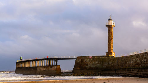 Lighthouse by pier at beach against cloudy sky