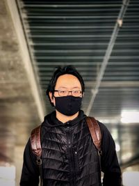 Portrait of young man in face mask standing against ceiling and wall.