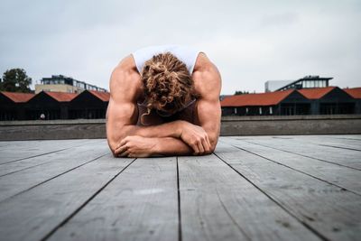 Muscular man lying on jetty by houses against sky