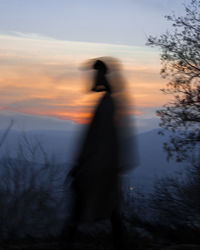 Rear view of woman standing by lake against sky during sunset