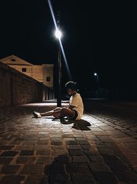 Teenage boy sitting on street by illuminated street light at night
