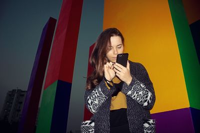 Young woman using mobile phone while standing against wall