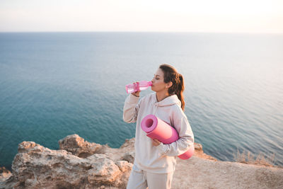 Pretty woman drinking water from pink bottle wear spotswear hoodie and pants hold yoga mat over sea