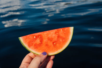 Hand holding up watermelon semi circle, half moon with lake, sea, water background on sunny day