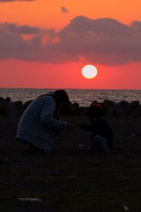 Silhouette people sitting on beach against sky during sunset