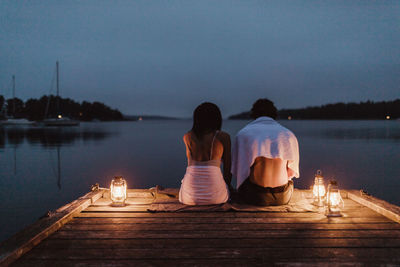 Rear view of couple sitting around illuminated lantern on pier by sea against sky