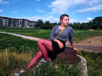 Full length of teenage girl with short hair looking away while sitting against sky at park during sunny day