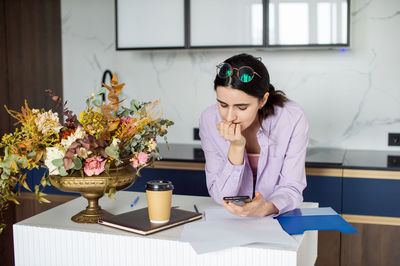 A cute girl is standing near a table with flowers, reading a message on her phone