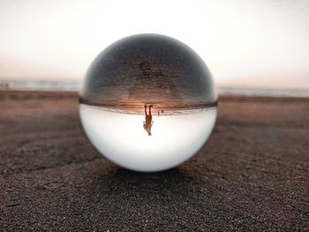 Reflection of man seen in crystal ball at beach against sky during sunset