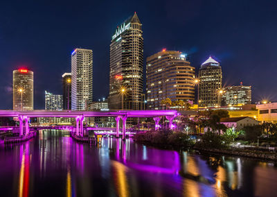 Illuminated purple bridge over river in city against sky