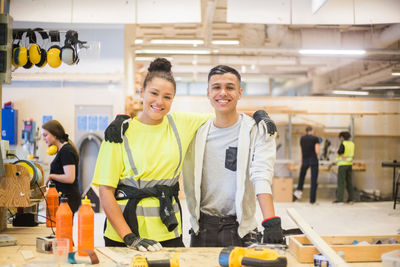 Portrait of smiling female trainee standing with arm around male coworker at workbench