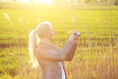 Side view of woman photographing with camera on field during sunset