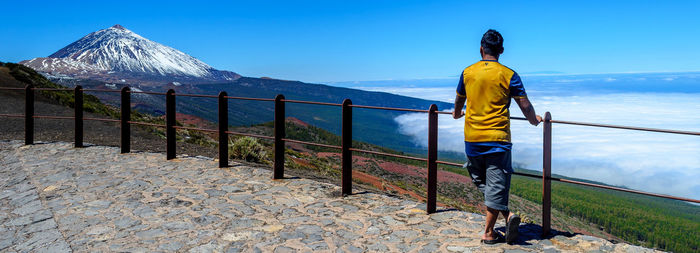 Rear view of man standing by railing on mountain by sea against sky