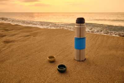 Vacuum flask and tea cups on a sandy beach. rising sun and sea at background. tea drinking outside