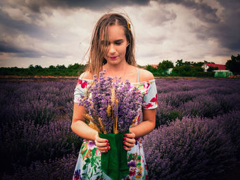 Woman holding lavender flowers while standing on field against cloudy sky