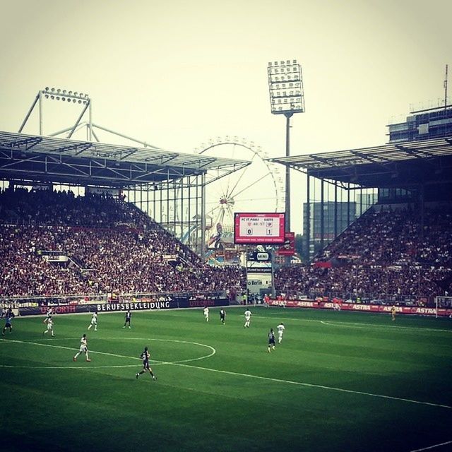 grass, sport, large group of people, built structure, architecture, building exterior, green color, men, field, leisure activity, communication, grassy, clear sky, person, outdoors, sky, day, soccer, city